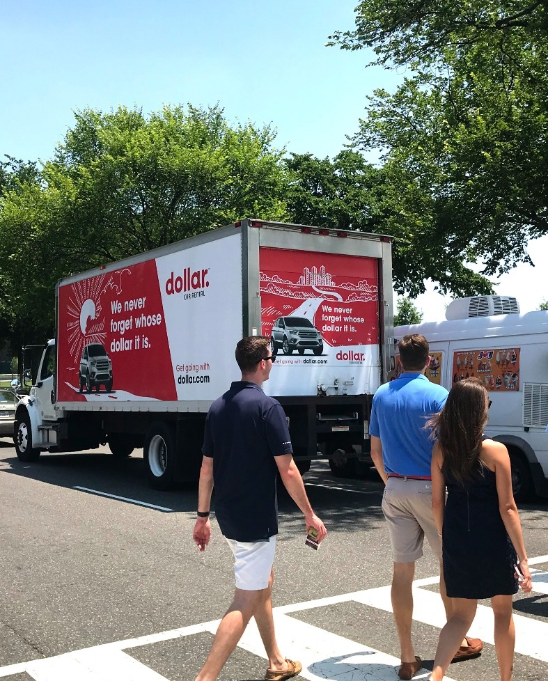 A real Dollar Car Rental-wrapped Advertising Wheels truck with the &lsquo;We never forget whose dollar it is&rsquo; campaign creative, with pedestrians on a crosswalk in view.